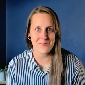 A person with long straight brown hair and light skin is smiling slightly at the camera. They are wearing a black and white vertical striped button-up shirt and have black ear gauges. The background is a solid dark blue wall with a small potted plant partially visible on the left side.