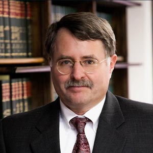 A middle-aged man with short dark hair, glasses, and a mustache wearing a dark suit and patterned tie, standing in front of wooden bookshelves filled with legal books.”