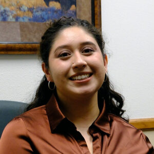 A portrait of a woman with shoulder-length dark hair, wearing a brown satin blouse and large hoop earrings. She has a bright smile, and the background shows a framed landscape painting.