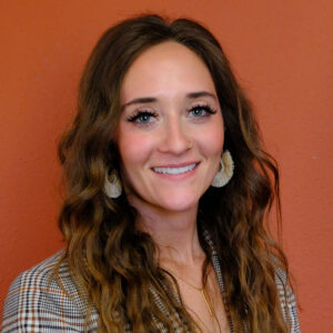 A smiling woman with long, wavy brown hair, wearing a checkered blazer and large earrings. The background is a warm reddish-orange color.