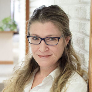 Woman with long, light brown hair and glasses, wearing a white blouse, smiling slightly while standing beside a white brick wall.